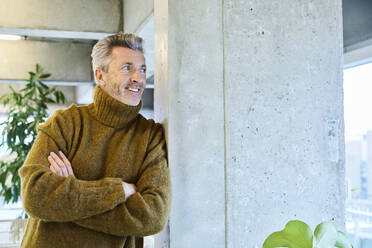 Man with arms crossed looking away while leaning on pillar at office - FMKF06861