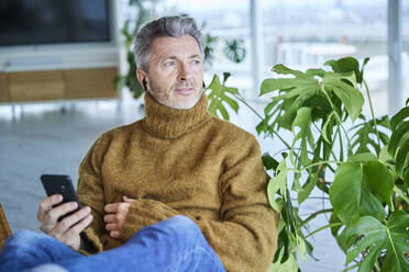 Älterer Mann mit In-Ear-Kopfhörern, der zu Hause sitzend ein Mobiltelefon benutzt - FMKF06872