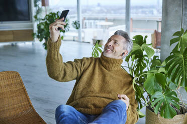 Lächelnder Mann, der ein Selfie mit seinem Mobiltelefon macht, während er zu Hause sitzt - FMKF06873