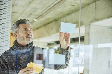 Male businessman putting sticky note while standing by glass wall at office - FMKF06876