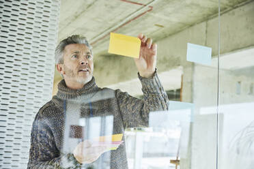 Mature businessman putting sticky note on glass wall while standing at office - FMKF06877