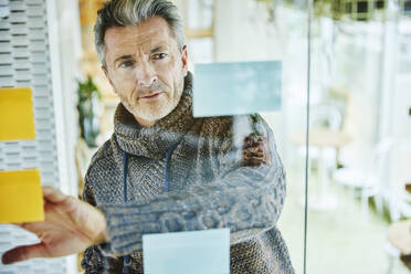 Mature businessman putting adhesive note on glass wall while standing at office - FMKF06878