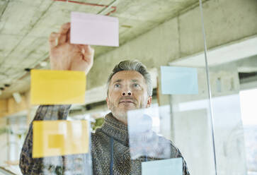 Male mature businessman putting note on glass wall while working at office - FMKF06879