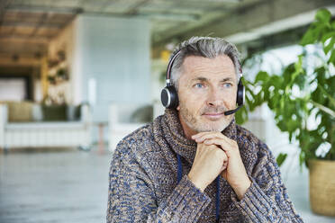 Businessman wearing headset sitting with hand on chin at office - FMKF06898