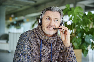 Älterer Geschäftsmann lächelt, während er über ein Headset im Büro spricht - FMKF06899