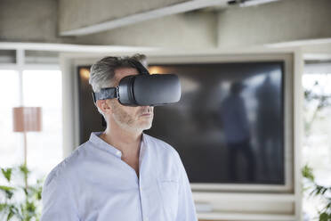 Mature businessman using virtual reality headset while standing at home - FMKF06944