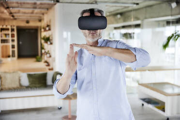 Male businessman wearing virtual reality headset gesturing while standing at home - FMKF06946