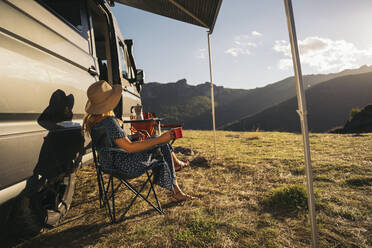 Frau auf Stuhl sitzend mit Kaffeetasse am Wohnmobil an einem sonnigen Tag - MPPF01326