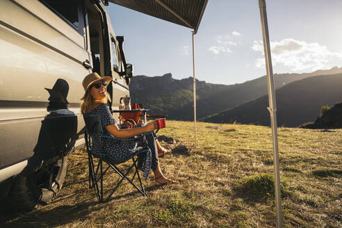Happy woman in sunglasses with coffee cup laughing while sitting by motor home during vacations - MPPF01327