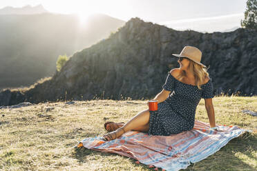Lächelnde Frau mit Kaffeetasse träumt beim Sitzen auf einer Picknickdecke an einem sonnigen Tag - MPPF01330