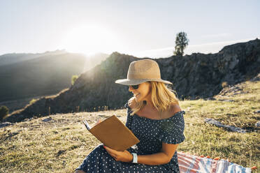 Glückliche Frau, die ein Buch liest, während sie auf einer Picknickdecke vor den Bergen an einem sonnigen Tag sitzt - MPPF01332