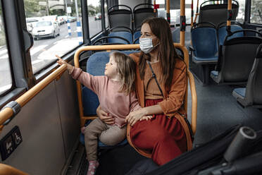 Mutter mit niedlicher Tochter, die auf das Fenster zeigt, während sie mit dem Bus während der COVID-19 reist - OGF00669