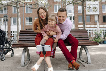 Happy family with one daughter talking selfie from mobile phone on tripod at park bench - OGF00701