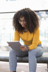 Lächelnde junge Frau mit lockigem Haar, die ein digitales Tablet benutzt, während sie auf einer Bank sitzt - PNAF00270