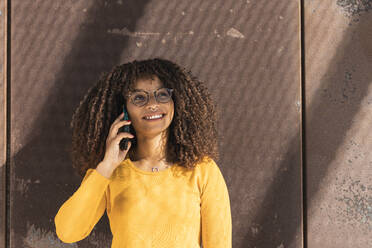 Lächelnde junge Frau, die an einem sonnigen Tag mit ihrem Handy an der Wand telefoniert - PNAF00276