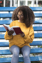 Modische junge Frau, die auf einer blauen Tribüne sitzt und ein Buch liest - PNAF00282