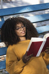 Smiling young woman reading book while lying on blue bleachers - PNAF00285