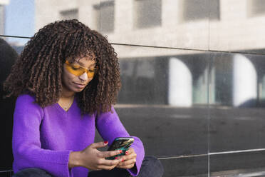 Afro-Frau mit Sonnenbrille und Smartphone an der Wand - PNAF00289