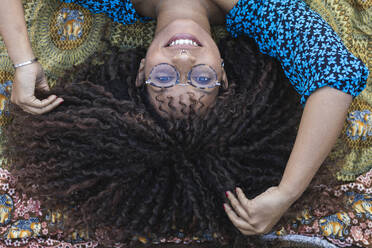 Lächelnde Afro-Frau mit Hand im Haar auf einer Decke im öffentlichen Park liegend - PNAF00307