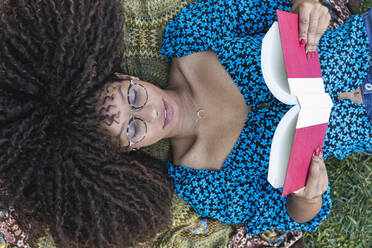 Afro young woman with curly hair reading book while lying down at public park - PNAF00308