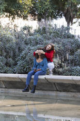 Smiling young woman covering eyes of girlfriend while sitting on retaining wall in public park - PNAF00309