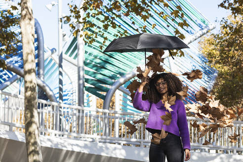 Afro junge Frau mit Regenschirm steht unter trockenen fallenden Blättern im Park - PNAF00318