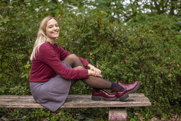 Smiling blond woman sitting on bench by plants in park during autumn - BFRF02336