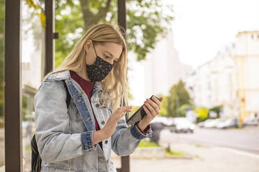 Woman using mobile phone at bus stop in city during COVID-19 - BFRF02348