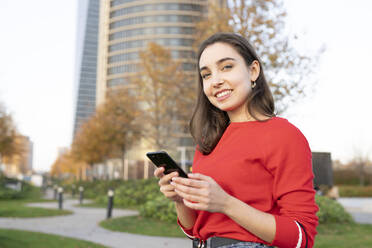 Smiling woman with mobile phone at park in city - JCCMF00231