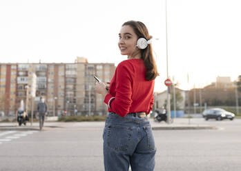 Woman with listening music through headphones while standing on street in city - JCCMF00233