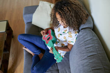 Smiling young woman with curly hair practicing to play ukulele in living room - KIJF03442