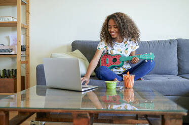 Smiling woman learning to play ukulele from laptop in living room - KIJF03445