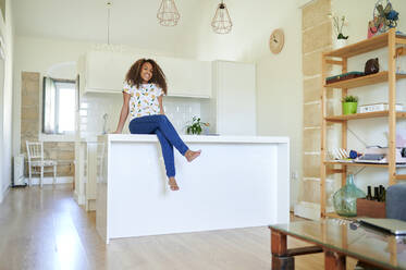 Lächelnde junge Afro-Frau, die zu Hause auf dem Küchentisch sitzt - KIJF03452