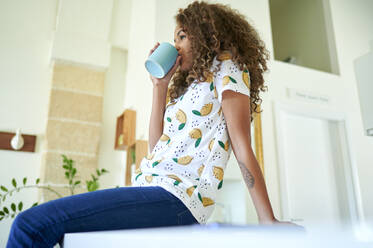 Afro young woman with curly hair drinking coffee while sitting on kitchen counter at home - KIJF03455