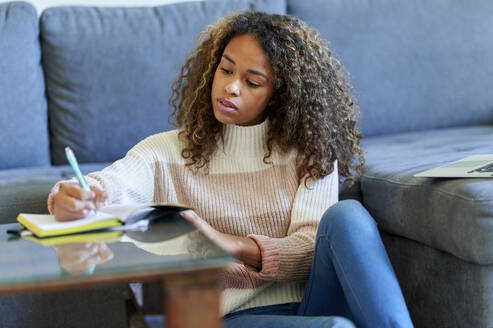 Junge Afro-Frau mit Laptop, die zu Hause in einen Notizblock schreibt - KIJF03483