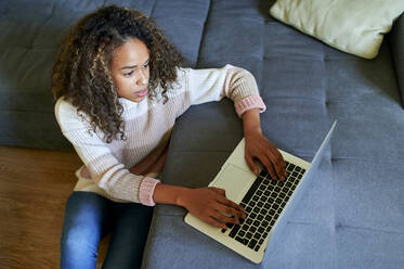 Junge Frau mit lockigem Haar benutzt Laptop auf Sofa im Wohnzimmer zu Hause - KIJF03488