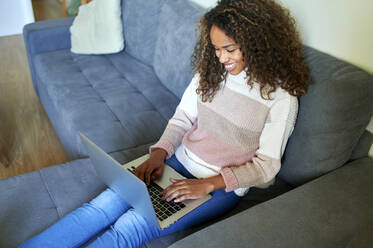 Smiling young woman using laptop while sitting on sofa at home - KIJF03489