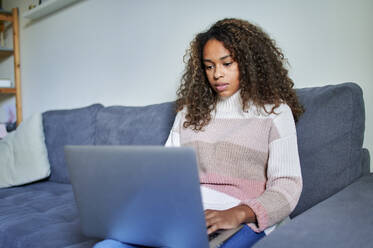 Young afro woman using laptop with concentration on sofa at home - KIJF03490
