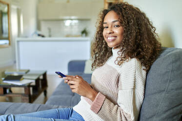 Smiling young woman with smart phone on sofa at home - KIJF03493