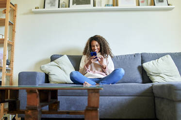Junge Afro-Frau lächelt, während sie auf dem Sofa im Wohnzimmer ein Mobiltelefon benutzt - KIJF03494