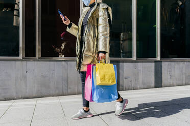 Junge Frau mit Gesichtsschutz, die Einkaufstüten trägt, während sie ein Mobiltelefon benutzt und auf einem Fußweg geht - MARF00038
