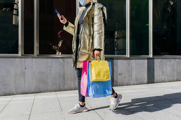 Young woman with protective face mask carrying shopping bags while using mobile phone walking on footpath - MARF00038