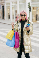Lächelnde Frau, die Einkaufstüten trägt, während sie ein Mobiltelefon benutzt, in der Stadt stehend - MARF00046