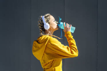 Sportswoman wearing headphones drinking water while standing against gray wall - PGF00253