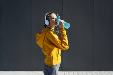 Weibliche Sportlerin mit Kopfhörern, die Wasser trinkt, während sie an einer grauen Wand steht - PGF00255