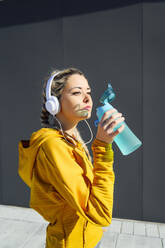 Sportler mit Kopfhörern, der Wasser trinkt, während er an einer grauen Wand steht - PGF00256