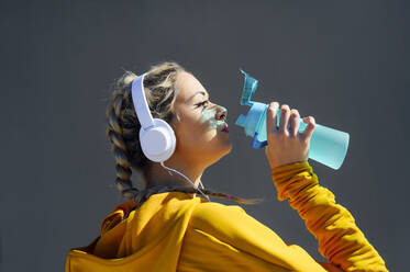 Junger Sportler mit Kopfhörern, der Wasser trinkt, während er an einer grauen Wand steht - PGF00257