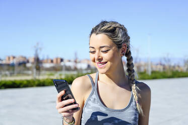 Sportlerin lächelt, während sie auf dem Gehweg stehend ein Mobiltelefon benutzt - PGF00279
