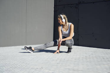 Young athlete wearing headphones stretching while crouching on footpath - PGF00309