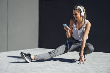 Smiling sportswoman wearing headphones using mobile phone while crouching on footpath - PGF00310